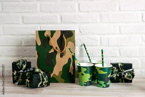 Party set with camouflage glasses, straws, shopping bag and gifts against the white brick wall. Copy space for text. photo