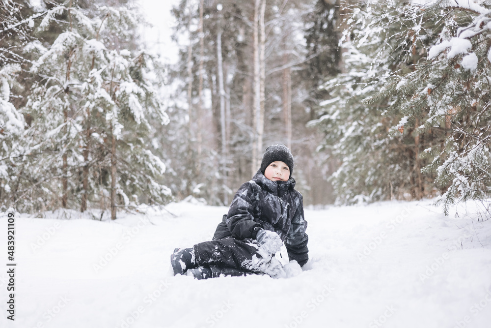 Happy teenager boy sitting on snow in winter forest. Child having fun outdoors. Joyful adolescent playing in snow at snowfall. Laughing smiling kid walking in winter park in cold weather