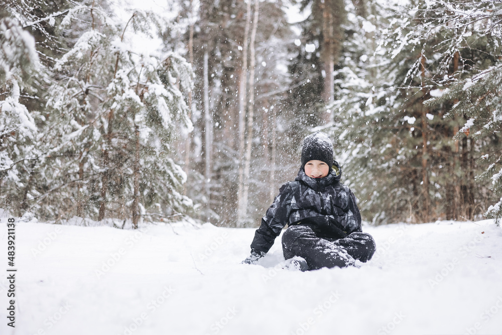 Happy teenager boy sitting on snow in winter forest. Child having fun outdoors. Joyful adolescent playing in snow at snowfall. Laughing smiling kid walking in winter park in cold weather