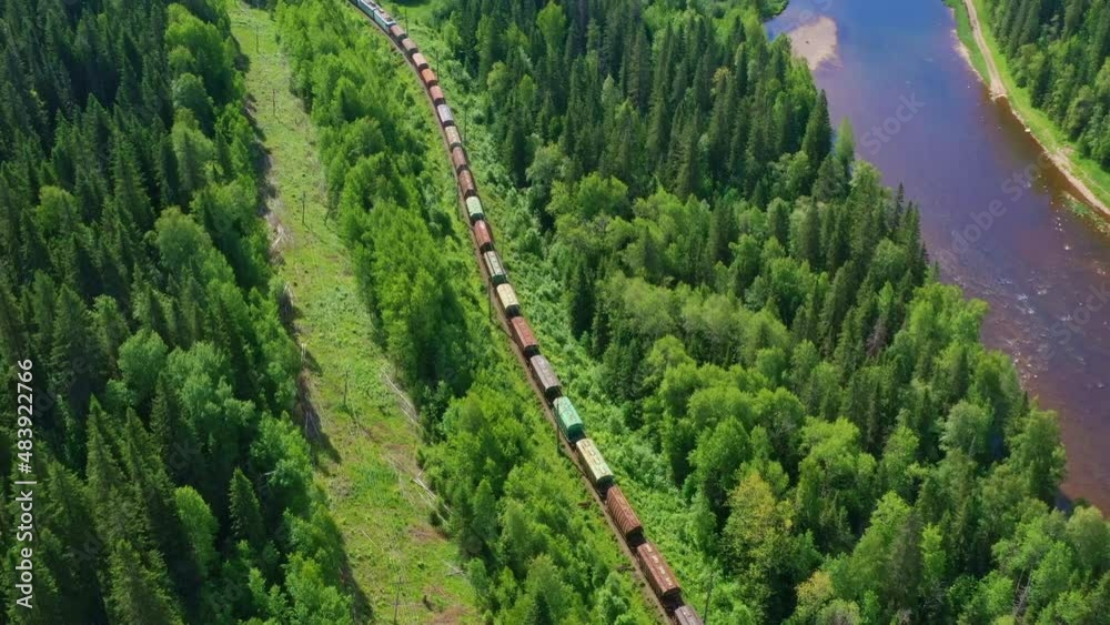 Aerial, top view of train moving along river trough forest. Carriages, cars ride on rails on summer day. Railroad transportation of goods taken by drone. Railway cargo, freight delivery services
