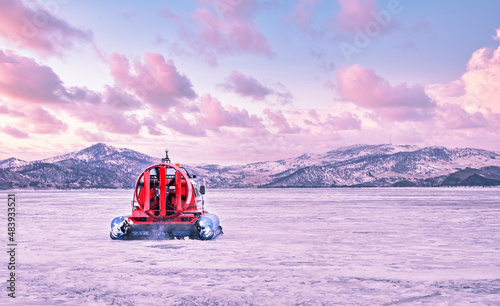 Hovercraft quickly glides over the surface of the transparent frozen Lake Baikal at sunset, winter pink landscape.