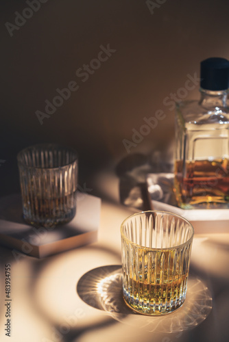 Whiskey with ice in glasses and bottle, beige background with hard light, shadows and sun glare, copy space