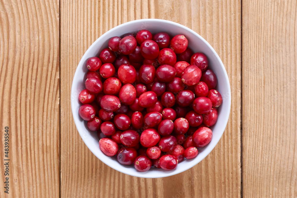 A white bowl with red ripe cranberries on a wooden background. Symmetrical top view image. A white bowl with red ripe cranberries on a wooden background. Symmetrical top view image.