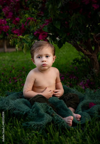 Baby boy is sitting in garden on the grass under the bush and lookin seriously