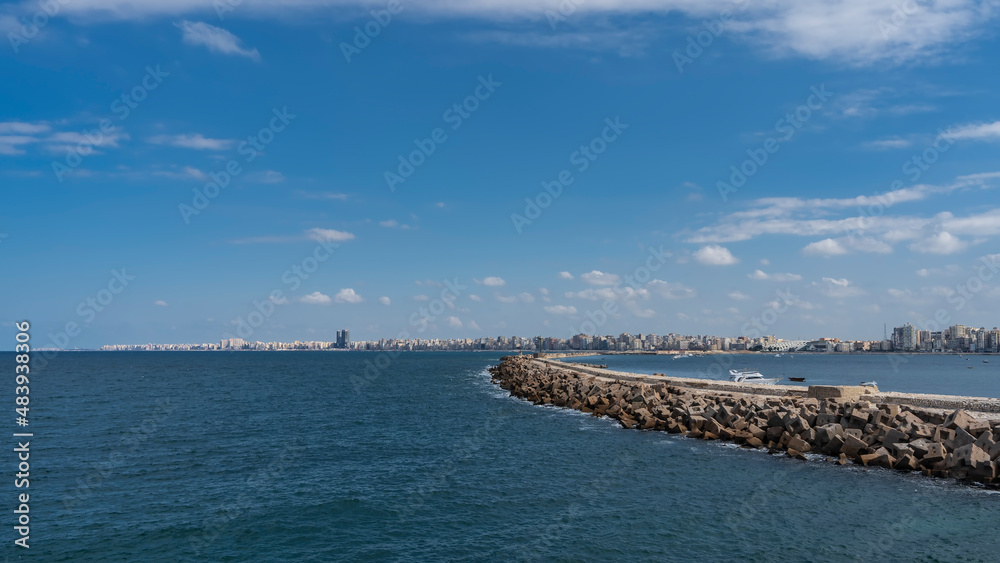 Seascape in Alexandria. A stone pier in the Mediterranean Sea, yachts are visible. In the distance, on the horizon, there is an embankment with city buildings. Blue sky with clouds. Egypt
