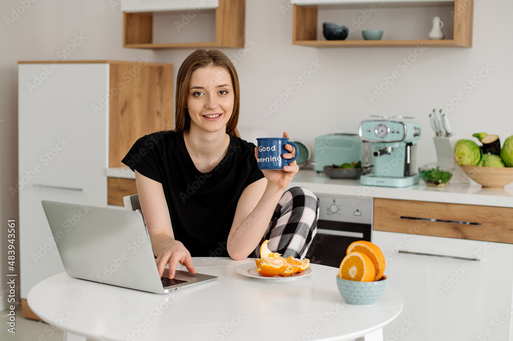 Beautiful young woman in pajamas working with laptop in the kitchen in the morning Beautiful young woman in pajamas working with laptop in the kitchen in the morning