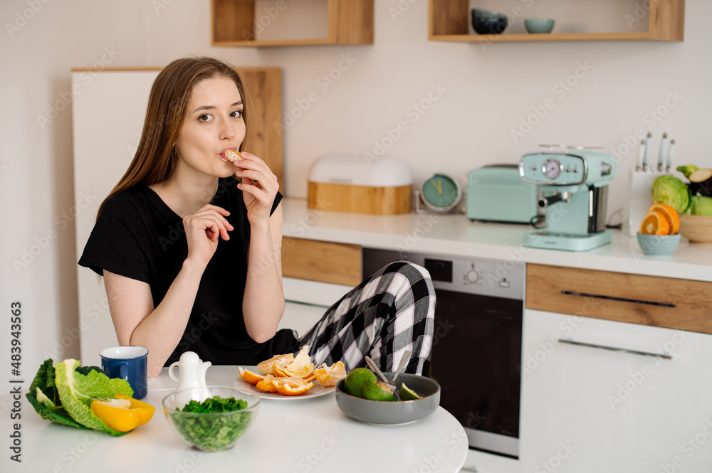 Young beautiful vegetarian girl dressed in pajamas eating fruits and vegetables for breakfast at home in the kitchen Young beautiful vegetarian girl dressed in pajamas eating fruits and vegetables for breakfast at home in the kitchen