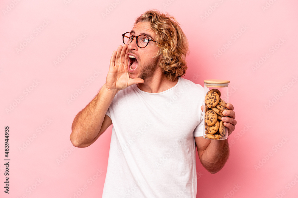Young caucasian man holding cookies jar isolated on pink background shouting and holding palm near opened mouth. Young caucasian man holding cookies jar isolated on pink background shouting and holding palm near opened mouth.