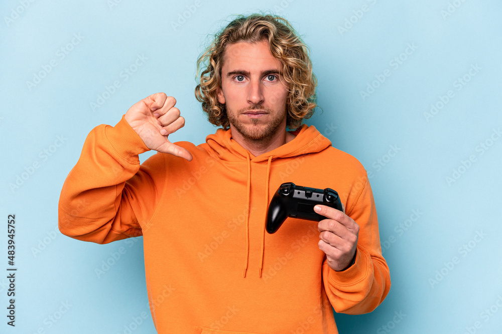 Young caucasian man playing with a video game controller isolated on blue background showing a dislike gesture, thumbs down. Disagreement concept. Young caucasian man playing with a video game controller isolated on blue background showing a dislike gesture, thumbs down. Disagreement concept.