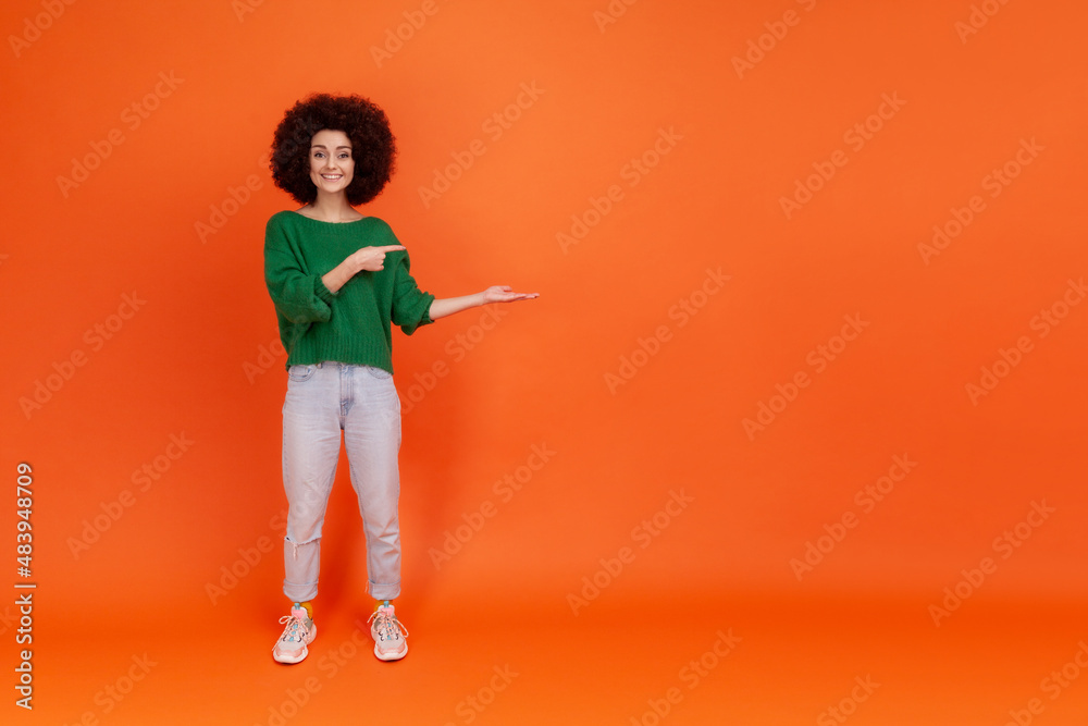 Full length of friendly smiling woman with Afro hairstyle wearing green casual style sweater presenting copy space for advertisement or promotion. Indoor studio shot isolated on orange background. Full length of friendly smiling woman with Afro hairstyle wearing green casual style sweater presenting copy space for advertisement or promotion. Indoor studio shot isolated on orange background.