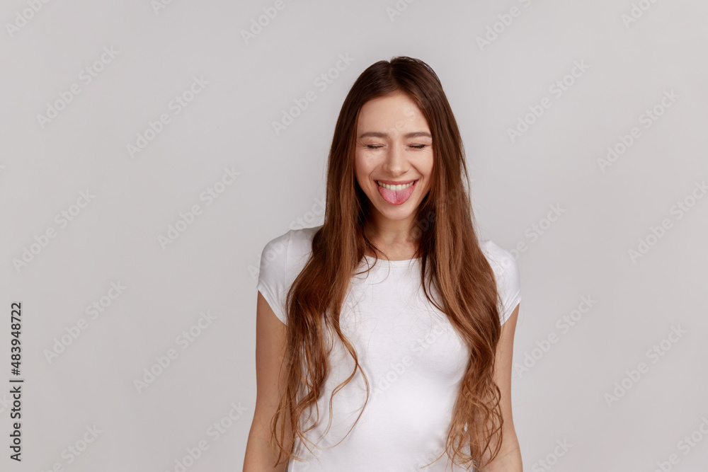 Portrait of positive playful woman standing with closed eyes and tongue out, demonstrating childish behavior, wearing white T-shirt. Indoor studio shot isolated on gray background. Portrait of positive playful woman standing with closed eyes and tongue out, demonstrating childish behavior, wearing white T-shirt. Indoor studio shot isolated on gray background.