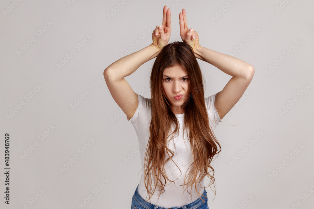 Fototapeta premium Portrait of aggressive bully woman with dark hair showing bull horns gesture over head, frowning as before attack, wearing white T-shirt. Indoor studio shot isolated on gray background.