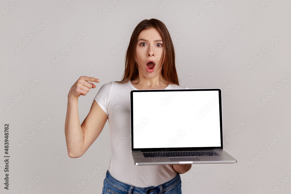 Shocked woman with dark hair pointing at white empty display of her portable computer, copy space for advertisement, wearing white T-shirt. Indoor studio shot isolated on gray background. Shocked woman with dark hair pointing at white empty display of her portable computer, copy space for advertisement, wearing white T-shirt. Indoor studio shot isolated on gray background.