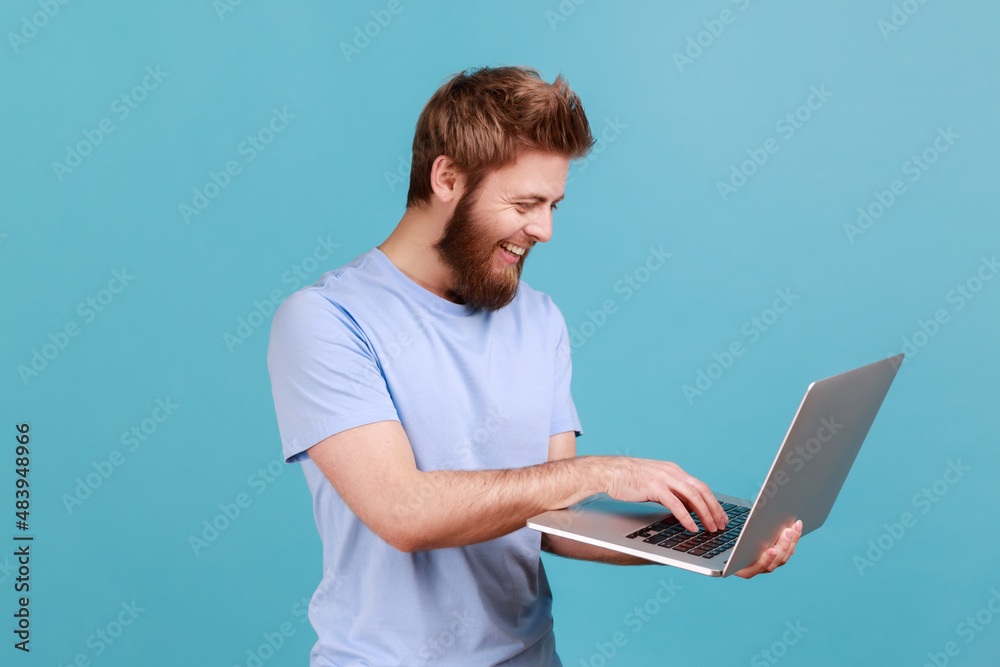 Portrait of smiling bearded man holding laptop in hand and typing, blogger making posts in social networks, chatting with followers. Indoor studio shot isolated on blue background.