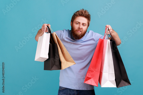 Portrait of tiered exhausted handsome bearded man raised arms with shopping bags,looking at camera, feels bored after going in mall. Indoor studio shot isolated on blue background. photo