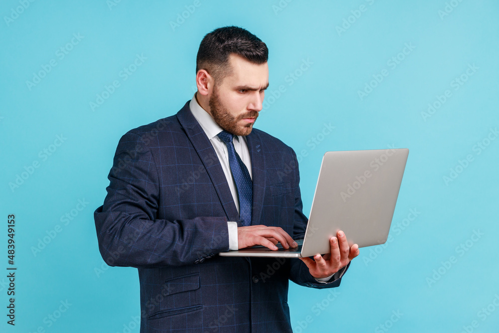 Young adult concentrated wearing official style suit holding laptop in hands, working on computer, looking at display with serious expression. Indoor studio shot isolated on blue background.