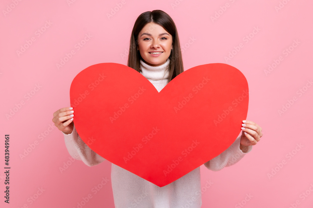 Portrait of young romantic female holding big red hear, expressing love, celebrating Valentines day, wearing white casual style sweater. Indoor studio shot isolated on pink background. Portrait of young romantic female holding big red hear, expressing love, celebrating Valentines day, wearing white casual style sweater. Indoor studio shot isolated on pink background.
