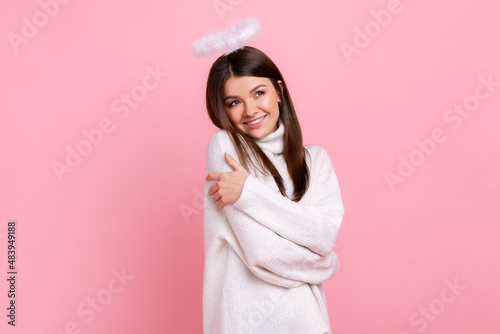 Принти на полотні Charming young adult brunette angelic woman with nimb over head, looking away and hugging herself, wearing white casual style sweater