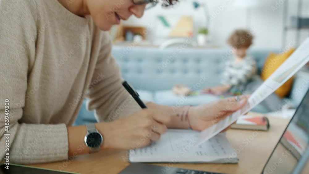 Serious freelancer working at home writing taking notes while son child playing in background. Motherhood and remote work concept. Serious freelancer working at home writing taking notes while son child playing in background. Motherhood and remote work concept.