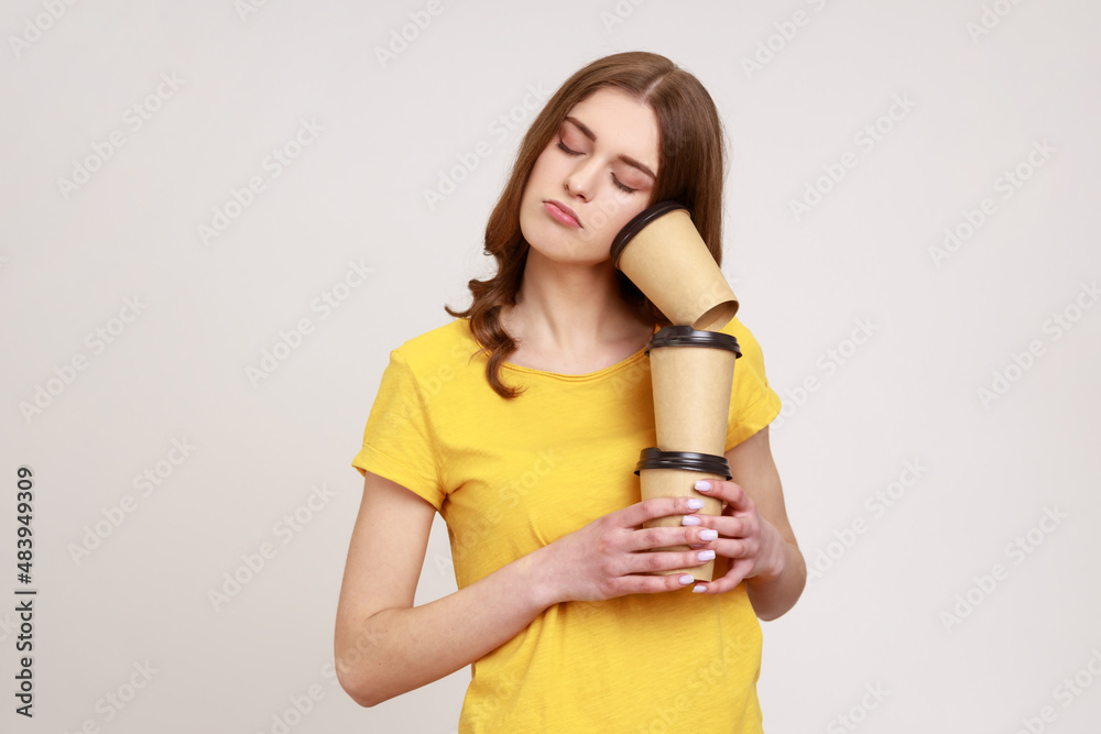 Tired sleepy young woman with brown hair in yellow T-shirt lying on coffee cups and sleeping, feeling exhausted bored after stressful night shift. Indoor studio shot isolated on gray background.