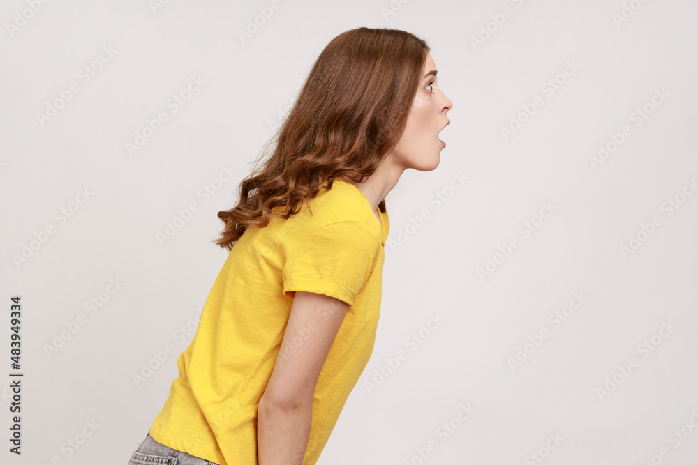 Side view of shocked beautiful young girl with brown wavy hair in yellow T-shirt standing and with surprised face, hearing shocking news. Indoor studio shot isolated on gray background. Side view of shocked beautiful young girl with brown wavy hair in yellow T-shirt standing and with surprised face, hearing shocking news. Indoor studio shot isolated on gray background.