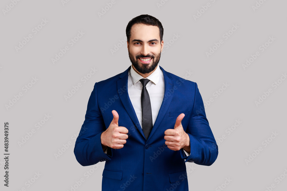 Portrait of happy bearded businessman showing thumbs up and smiling excitedly, enjoying excellent result, wearing official style suit. Indoor studio shot isolated on gray background. Portrait of happy bearded businessman showing thumbs up and smiling excitedly, enjoying excellent result, wearing official style suit. Indoor studio shot isolated on gray background.