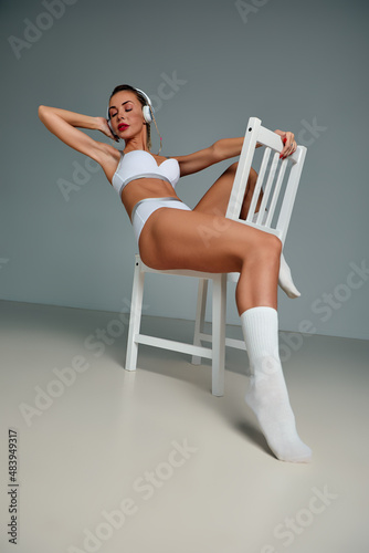 Banner of young happy girl in white swimsuit listening to music in white chair, wearing wireless headhones, isolated on gray background with copy space in studio photo