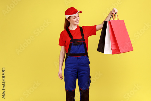 Portrait of smiling delivery woman standing with shopping bags in hands, waiting client to deliver parcels, wearing overalls and red cap. Indoor studio shot isolated on yellow background.