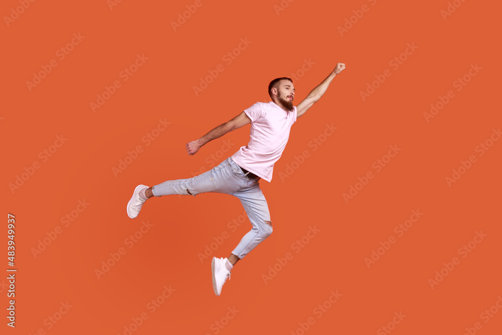 Portrait of motivated confident bearded man flying up in air as superhero with raised hand, feeling superpower, wearing pink T-shirt. Indoor studio shot isolated on orange background. Portrait of motivated confident bearded man flying up in air as superhero with raised hand, feeling superpower, wearing pink T-shirt. Indoor studio shot isolated on orange background.