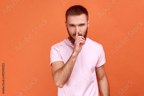 Portrait of bearded man holding finger on lips making hush silence gesture, asking to keep secret, don't speak, wearing pink T-shirt. Indoor studio shot isolated on orange background.