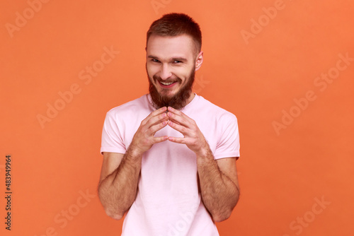Portrait of cunning handsome bearded man with envy smile scheming and conspiring, thinking devious tricks and cheats, wearing pink T-shirt. Indoor studio shot isolated on orange background. photo