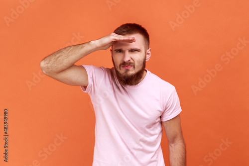 Portrait of bearded man looking far away at distance with hand over head, attentively searching for bright future, wearing pink T-shirt. Indoor studio shot isolated on orange background.