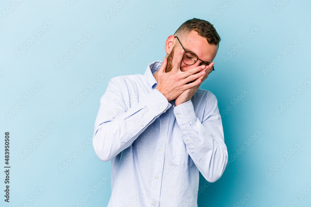 Young caucasian man isolated on blue background blink at the camera through fingers, embarrassed covering face. Young caucasian man isolated on blue background blink at the camera through fingers, embarrassed covering face.