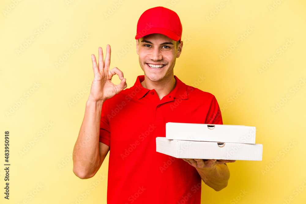 Young delivery caucasian man holding pizzas isolated on yellow background cheerful and confident showing ok gesture. Young delivery caucasian man holding pizzas isolated on yellow background cheerful and confident showing ok gesture.