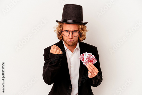 Young magician caucasian man holding magic cards isolated on white background showing fist to camera, aggressive facial expression.
