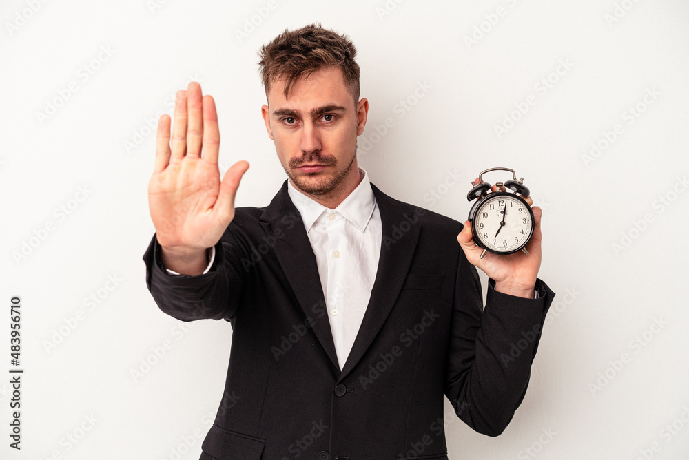 Young caucasian business man holding alarm clock isolated on white background standing with outstretched hand showing stop sign, preventing you. Young caucasian business man holding alarm clock isolated on white background standing with outstretched hand showing stop sign, preventing you.