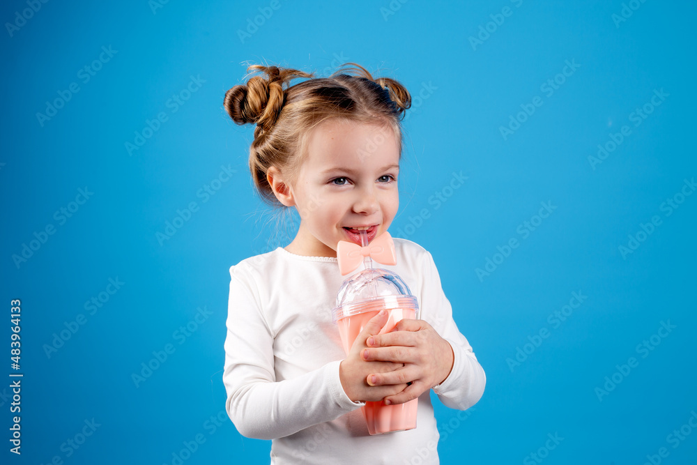 cute little girl with a funny hairstyle in a white T-shirt is holding a smoothie glass in her hands. blue background. useful products for children. space for text. High quality photo cute little girl with a funny hairstyle in a white T-shirt is holding a smoothie glass in her hands. blue background. useful products for children. space for text. High quality photo