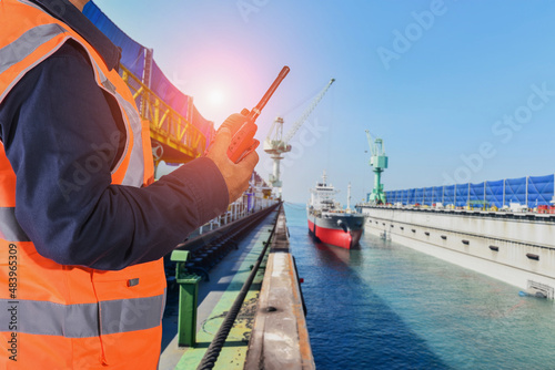 A man holding a walkie talke, or radio, wearing reflective shirt, controls the cargo ship into the dry dock for ship repair, maintenance in shipyard with copy space for texture. photo