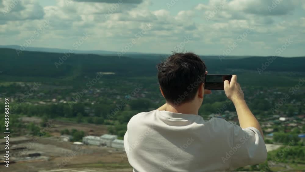 A young adult male takes a smartphone picture from the top of a large hill. A young adult male takes a smartphone picture from the top of a large hill.