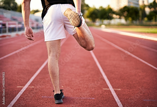 Young muscular athletic runner man stretching his leg on a running court in standing position before starting of running from back view - Jogging and flexibility concept