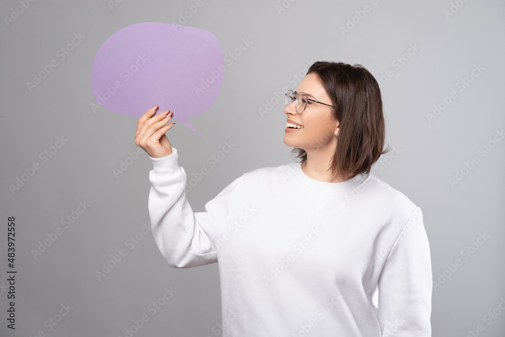 Cheerful young woman holding blank speech bubble over grey background. Cheerful young woman holding blank speech bubble over grey background.