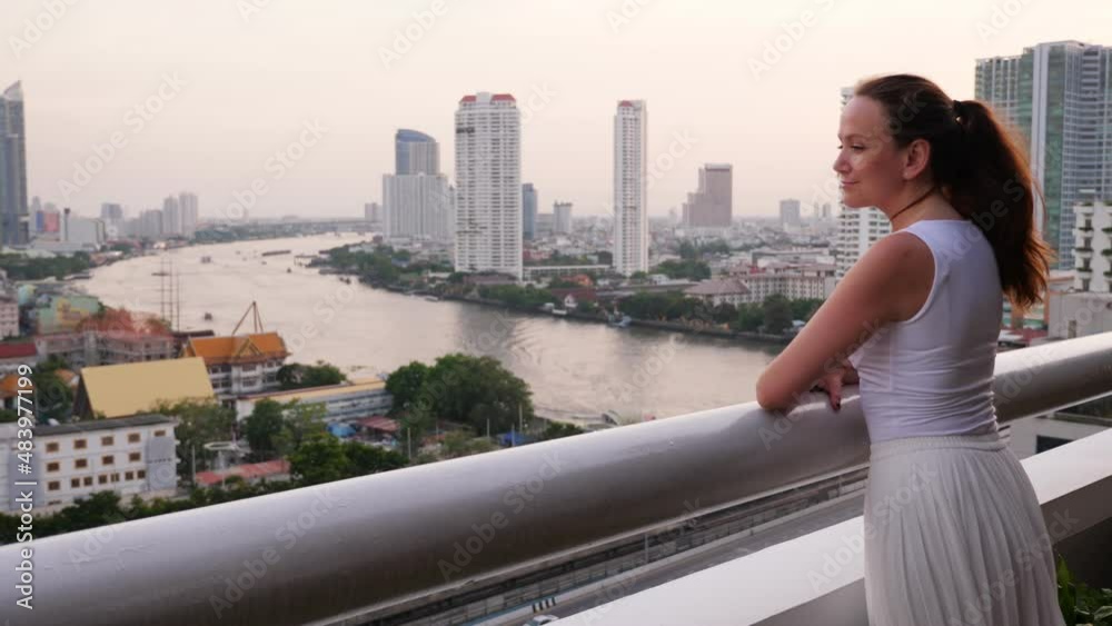 Tourist woman admiring a beautiful view of the city from a high floor, leaning on the railing and looking at Bangkok panorama. The Chao Phraya river and city buildings are visible in background Tourist woman admiring a beautiful view of the city from a high floor, leaning on the railing and looking at Bangkok panorama. The Chao Phraya river and city buildings are visible in background