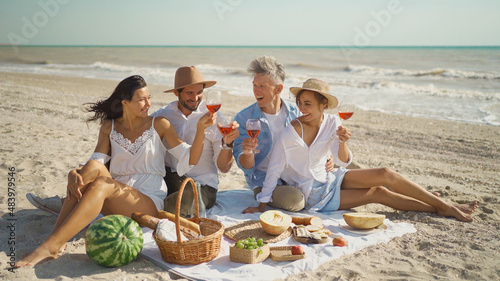 Happy group of friends enjoying beach picnic together, sharing conversation, drinking wine, celebrating weekend reunion gathering