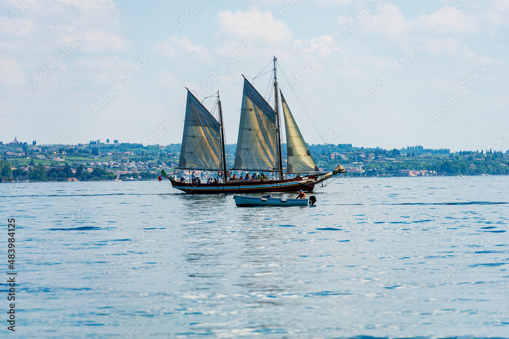Old wooden sailing ship and a motorboat in motion on the Lake Garda in front of the small town of Garda, tourist resort in Verona province, Veneto, Italy, southern Europe. Old wooden sailing ship and a motorboat in motion on the Lake Garda in front of the small town of Garda, tourist resort in Verona province, Veneto, Italy, southern Europe.