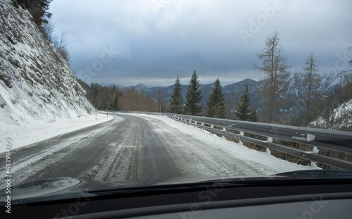 A shot on the move from behind the windshield of an electric car with snow-covered mountains. Cold cloudy winter day. POV first person view shot on a snowed mountain road. Selective focus.