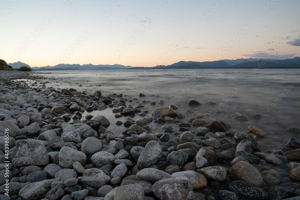 Long exposure shot of Nahuel Huapi lake and the Andes mountains in the background, at sunset. Beautiful blurred water effect, the rocky shore and dusk colors in the landscape. 