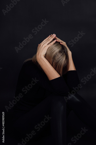 Caucasian woman in depression wearing black dress being on floor hiding face with arms in despair with black background. Mental and physical abuse. Rough patch.