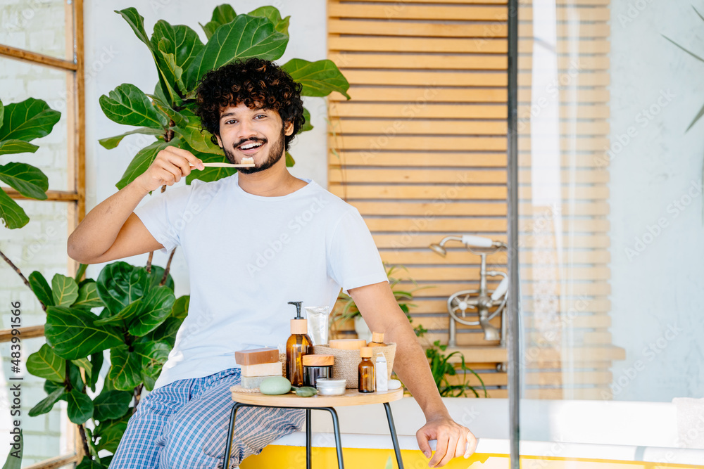 Happy bearded man sitting on bathtub smiling at camera holding wooden toothbrush with toothpaste. Eco-friendly beauty and cosmetic products. Zero waste and sustainable plastic free lifestyle. Happy bearded man sitting on bathtub smiling at camera holding wooden toothbrush with toothpaste. Eco-friendly beauty and cosmetic products. Zero waste and sustainable plastic free lifestyle.