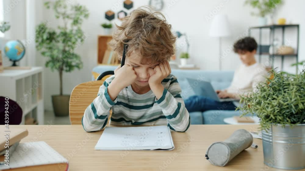 Unhappy child doing homework in apartment while young mother using laptop in background. Negative emotions and domestic education concept. Unhappy child doing homework in apartment while young mother using laptop in background. Negative emotions and domestic education concept.