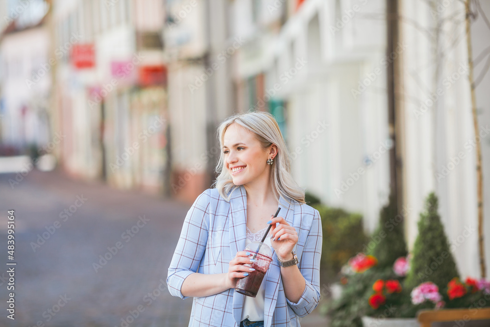 Stylish blonde in casual light clothes with a cocktail in her hands. Portrait of a beautiful fashionable woman on the street of a European city. Warm time. Shopping. Stylish blonde in casual light clothes with a cocktail in her hands. Portrait of a beautiful fashionable woman on the street of a European city. Warm time. Shopping.